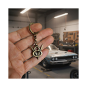 Hand hält V8 Schlüsselanhänger aus Messing vor einem Muscle‑Car in der Autowerkstatt.