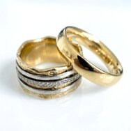 Zwei Trauringe in 585 Gold auf weißem Untergrund. Links ein breiter Ring mit organischer Wellenform, weißgold-farbenen Streifen und eingefassten Diamanten. Rechts ein klassischer, glänzender Goldring in schlichter gewölbter Form.